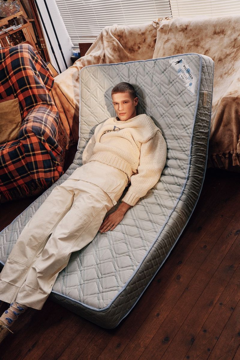 Adult male lying on a quilted mattress indoors, wearing cream sweater and pants, with a plaid chair in the background.