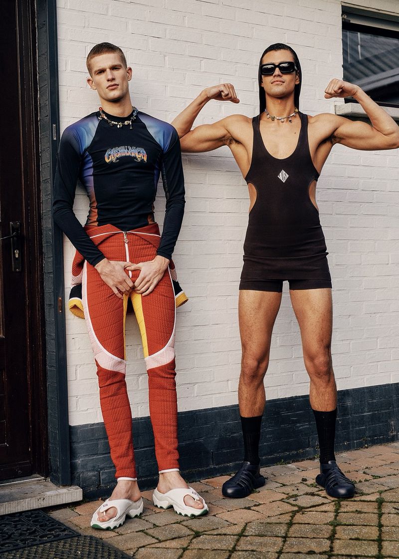 Two adult men pose in front of a white brick wall; one flexes, wearing a black one-piece, while the other wears athletic apparel.