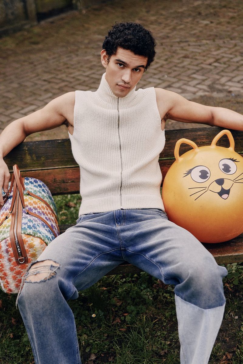 Young man sits on a park bench, wearing a knit vest, jeans with rips, and a colorful bag and toy next to him in daylight.