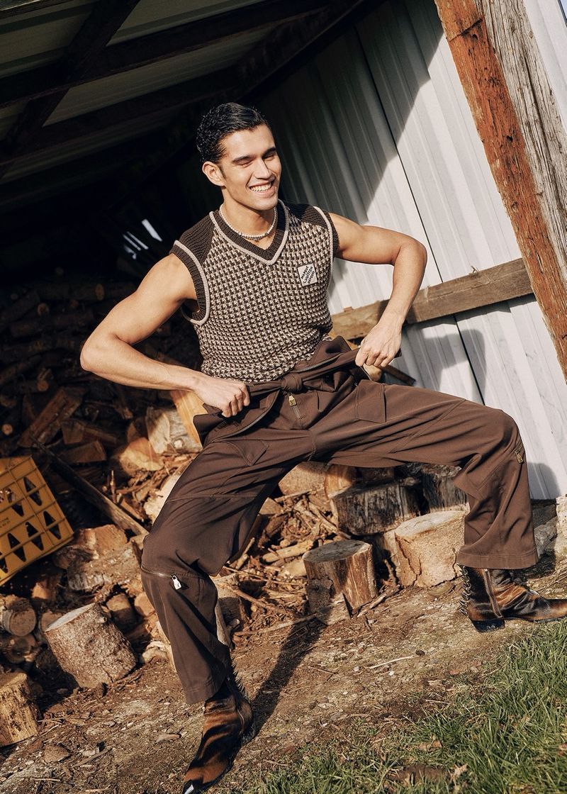 A young adult male with dark hair poses in front of a rustic wooden structure, wearing a patterned vest and brown work pants in outdoor setting.