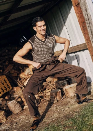 A young adult male with dark hair poses in front of a rustic wooden structure, wearing a patterned vest and brown work pants in outdoor setting.