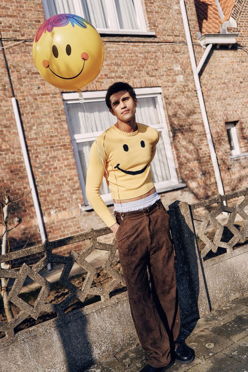 Stylish young adult male poses outdoors with a smiley-face balloon, wearing a matching yellow sweater and brown corduroy pants near brick house.