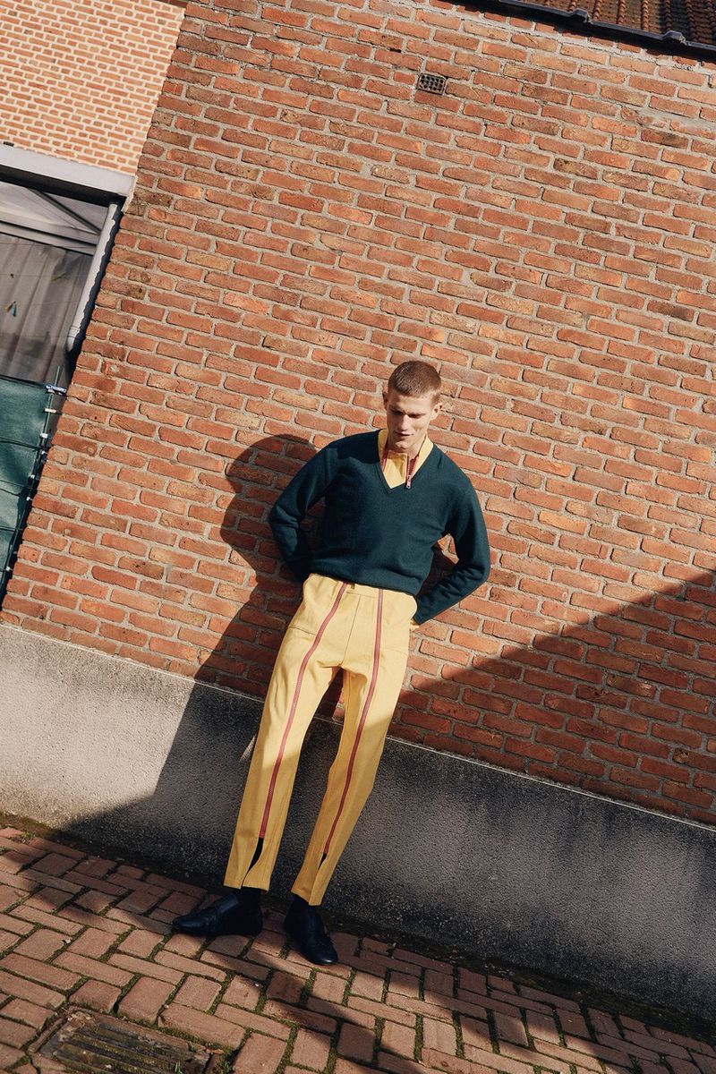 Fashion shot of a young adult man posing outdoors wearing a dark v-neck sweater, yellow pants, and black boots against a brick wall.