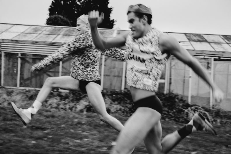 Two male athletes captured mid-run in black and white, wearing athletic gear, sneakers, and caps, with a greenhouse in the backdrop.
