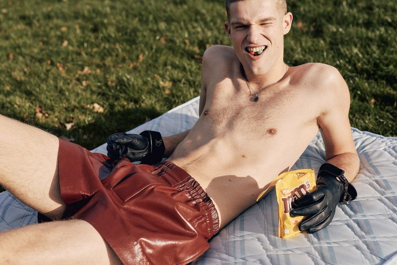 Shirtless man lies on a mattress in the grass wearing boxing gloves and holding a bag of M&Ms candy with a piece in his mouth.