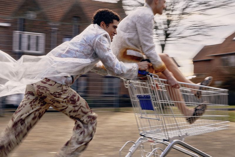 Two young adult men in a shopping cart being pushed outdoors. One man is pushing the cart, and the other is riding inside.