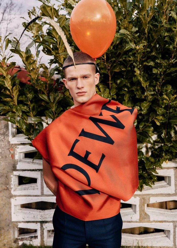Model wearing a structured orange top with large dark lettering outdoors with an orange balloon and feather accessory.