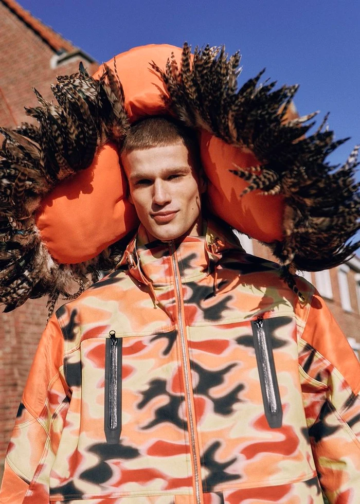 Model wearing orange camo jacket with feathered hood against building exterior for L'Officiel Belgium