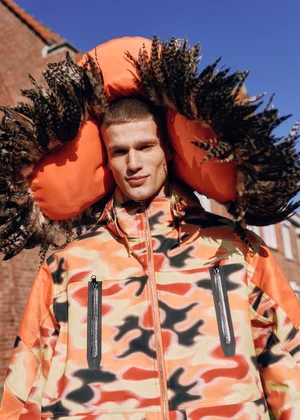 Model wearing orange camo jacket with feathered hood against building exterior for L'Officiel Belgium