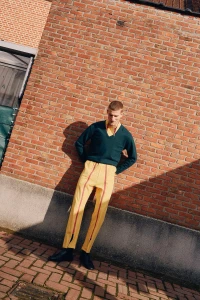Model leaning against a red brick wall wearing a dark green V-neck sweater and yellow trousers