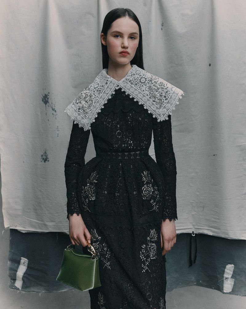A woman in a black lace dress with a white lace collar holds a small green purse against a muted studio backdrop. Fashion editorial.