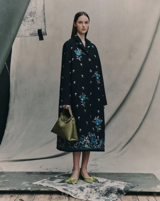 Fashionable young woman posing in a black floral coat with a green handbag and shoes against a neutral backdrop in a studio setting.