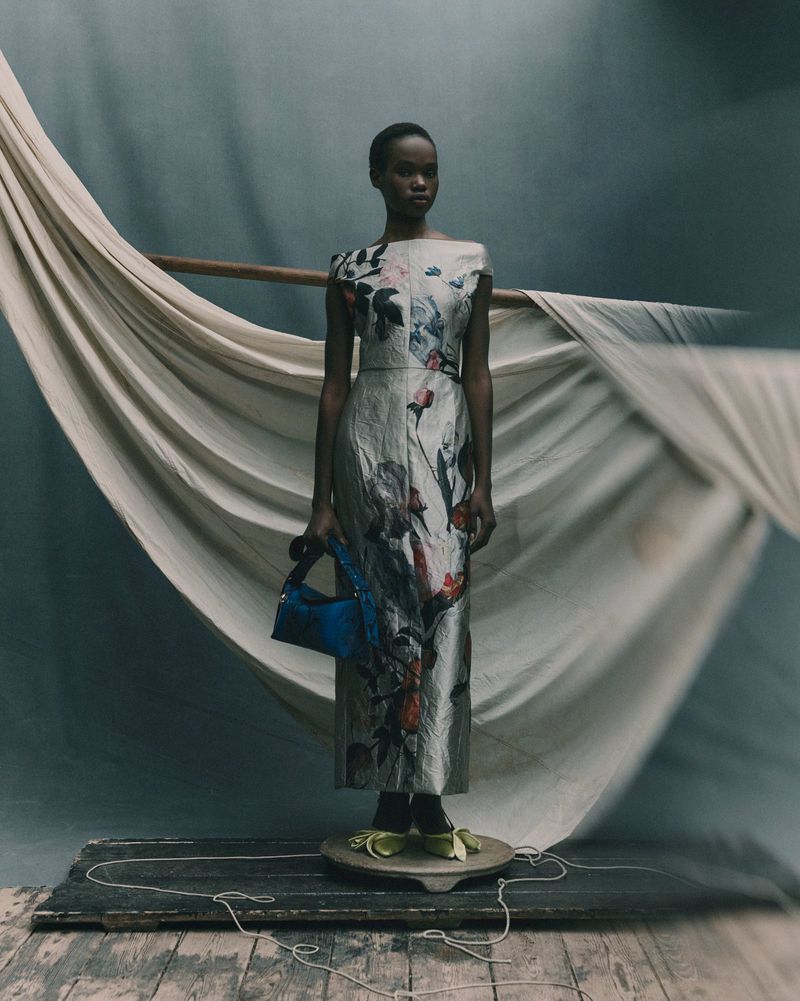 A woman stands in a silver floral dress with a blue bag on a wooden platform. The backdrop includes white fabric and a muted color palette.