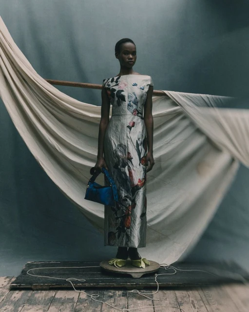 A woman stands in a silver floral dress with a blue bag on a wooden platform. The backdrop includes white fabric and a muted color palette.
