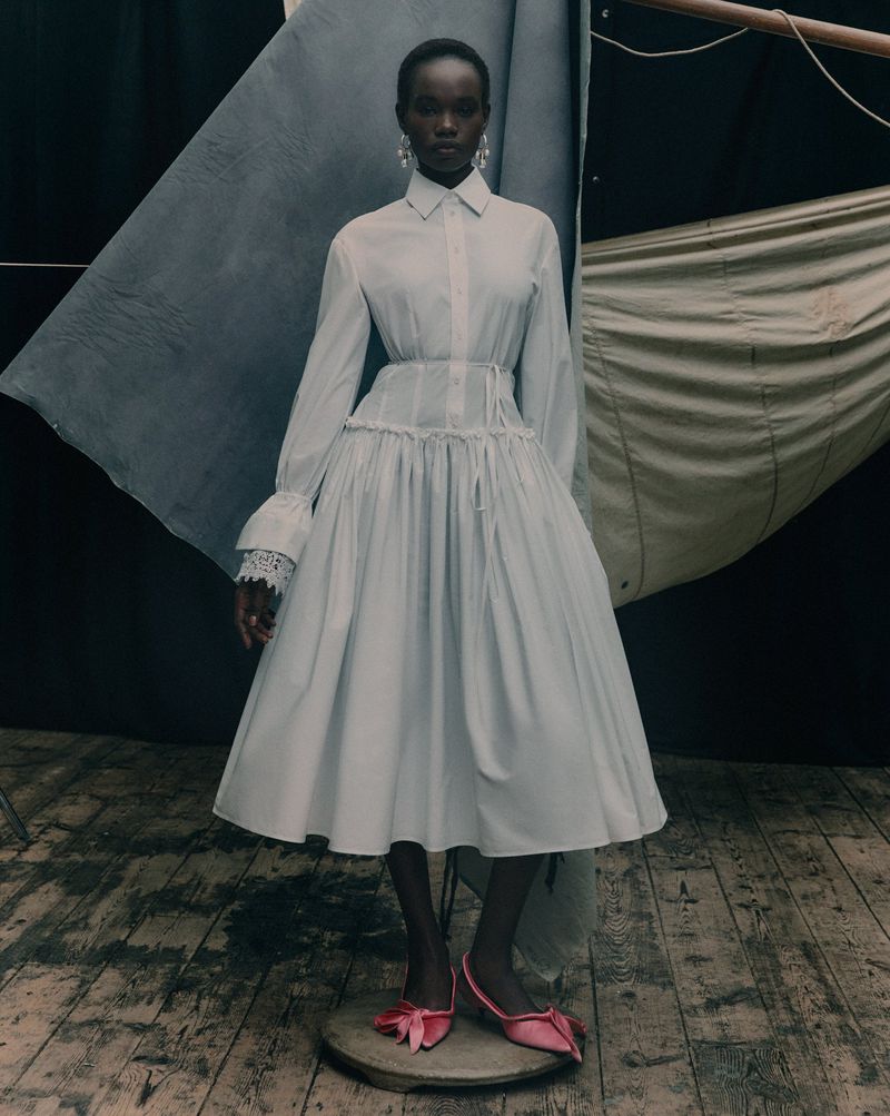A solo female fashion model in a white dress with lace sleeves and pink shoes poses against a backdrop of draped cloth in a studio.