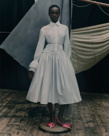 A solo female fashion model in a white dress with lace sleeves and pink shoes poses against a backdrop of draped cloth in a studio.