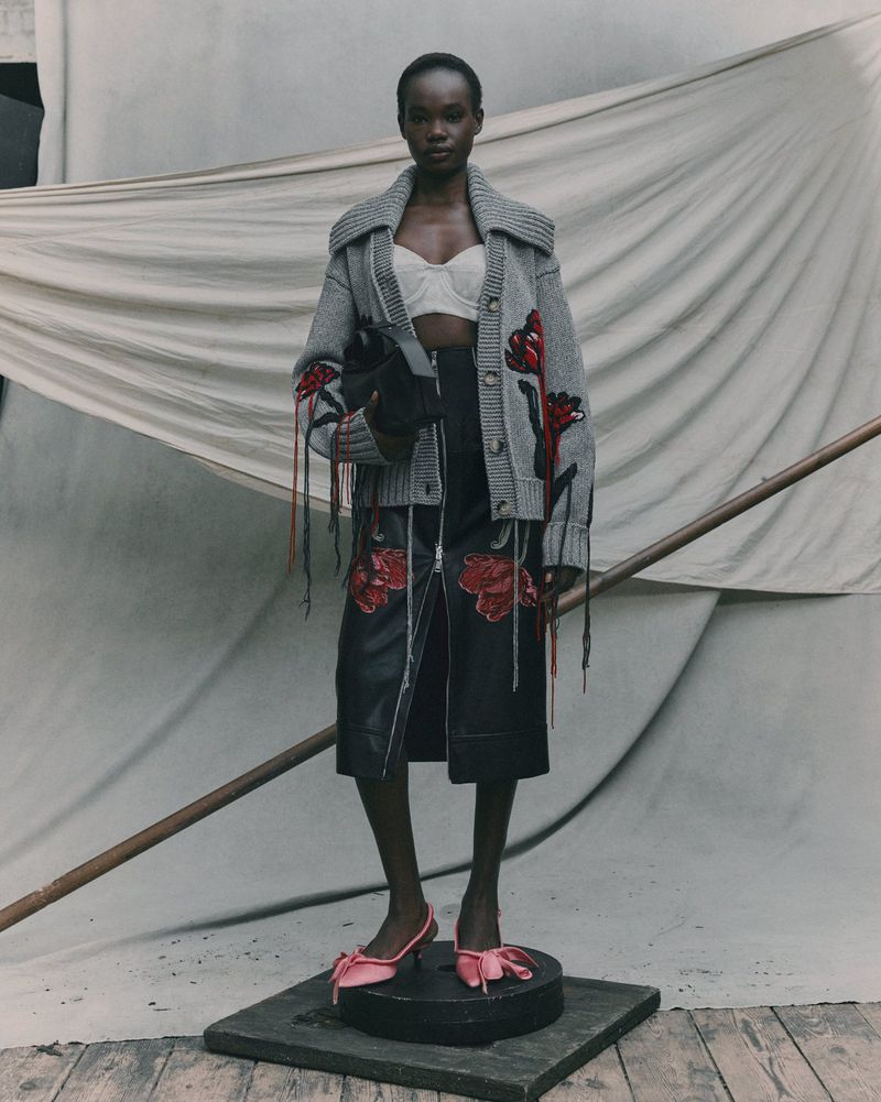 Fashion shot of a Black woman in a grey cardigan, black skirt with flower details, and pink bow heels, standing in a neutral studio.
