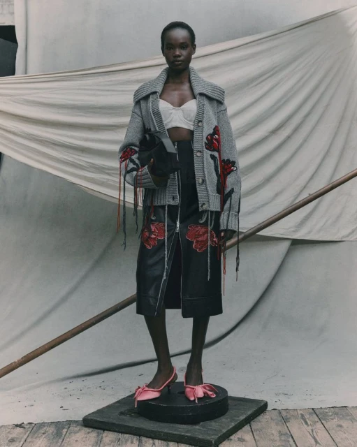Fashion shot of a Black woman in a grey cardigan, black skirt with flower details, and pink bow heels, standing in a neutral studio.