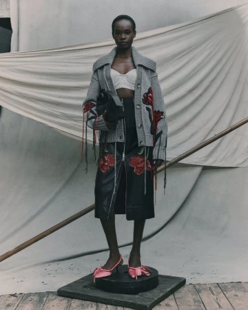 Fashion shot of a Black woman in a grey cardigan, black skirt with flower details, and pink bow heels, standing in a neutral studio.