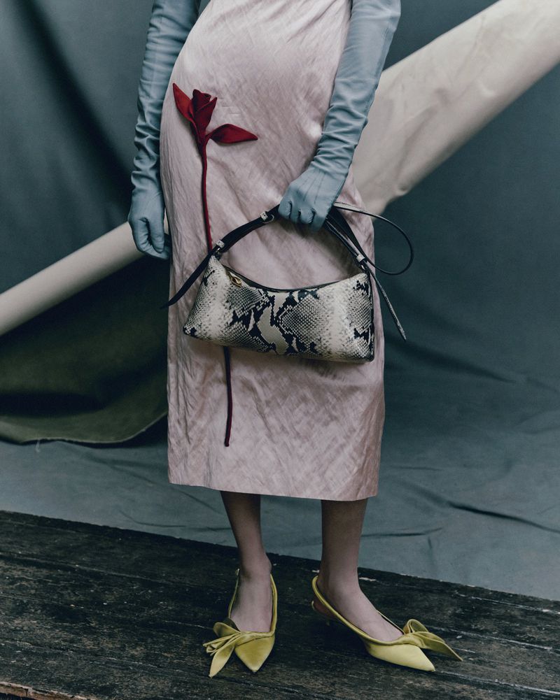 Fashion editorial shot featuring a woman in a pink dress holding a snakeskin purse, paired with yellow bow shoes and long gloves against a grey backdrop.