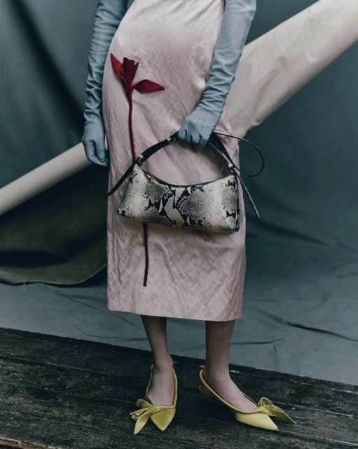 Fashion editorial shot featuring a woman in a pink dress holding a snakeskin purse, paired with yellow bow shoes and long gloves against a grey backdrop.