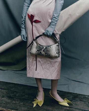 Fashion editorial shot featuring a woman in a pink dress holding a snakeskin purse, paired with yellow bow shoes and long gloves against a grey backdrop.