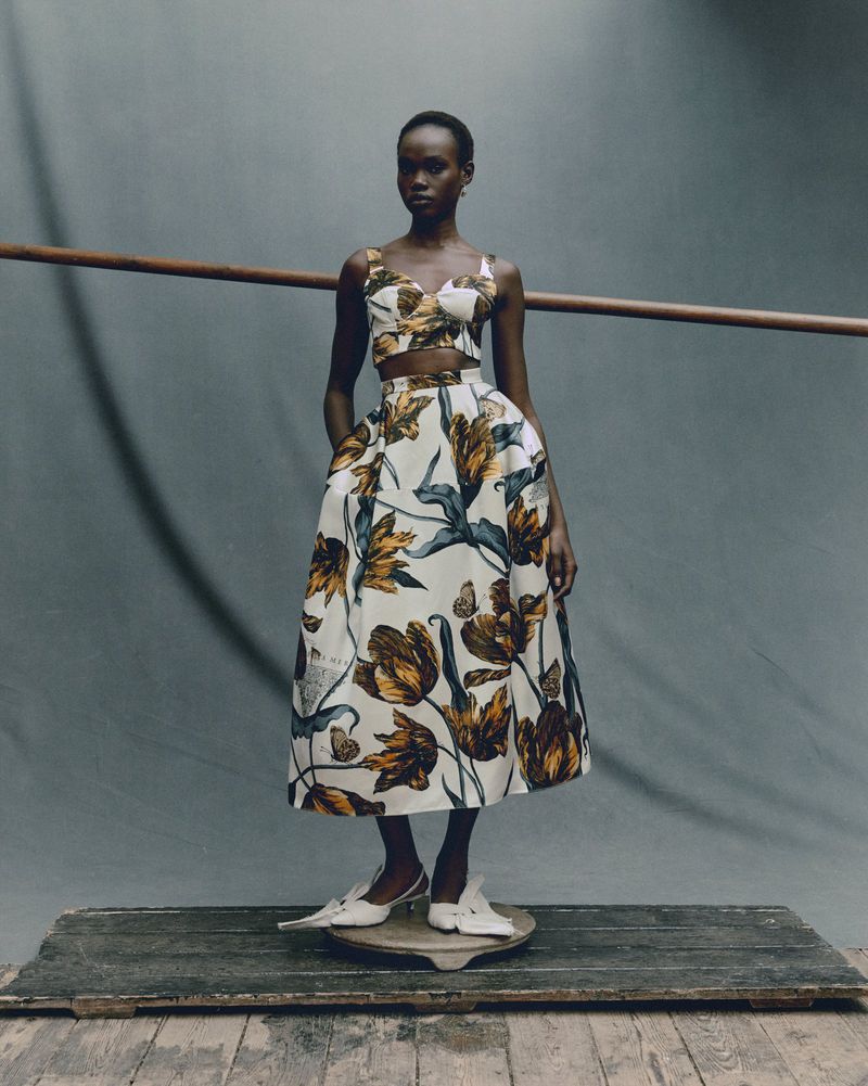 Elegant Black woman in a floral two-piece outfit poses in front of a gray background with a wooden rail and a platform stand.