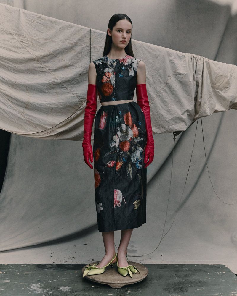 A woman stands wearing a floral print top and skirt, bright red opera gloves, and green heels, in a fashion photography studio setting.
