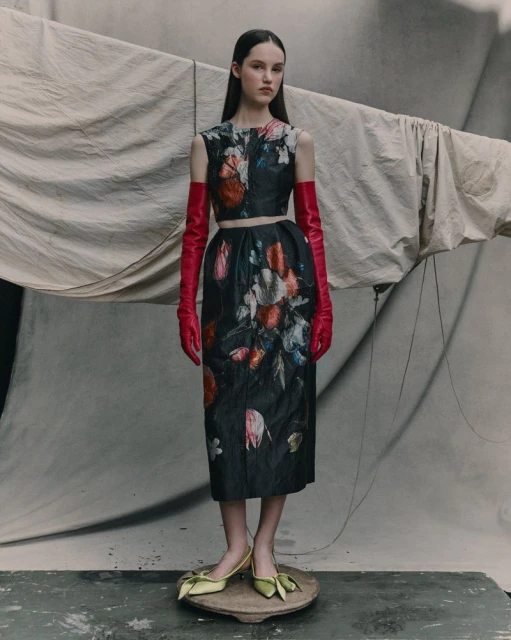 A woman stands wearing a floral print top and skirt, bright red opera gloves, and green heels, in a fashion photography studio setting.