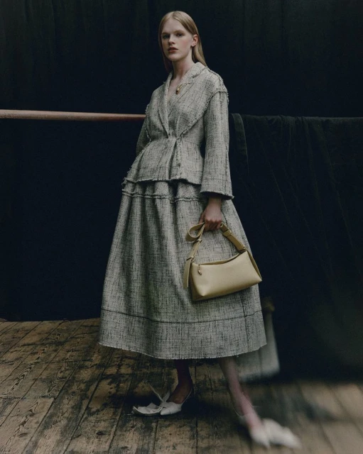 Fashion shot of woman in tweed skirt suit with purse in studio, spotlight on model, dark background, natural light from right.