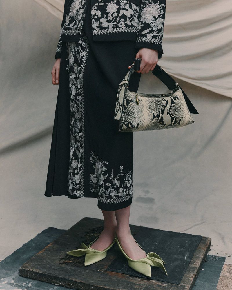 Fashionable woman in black skirt and jacket with floral embroidery, holding a snakeskin handbag, and wearing green bow-tie heels.