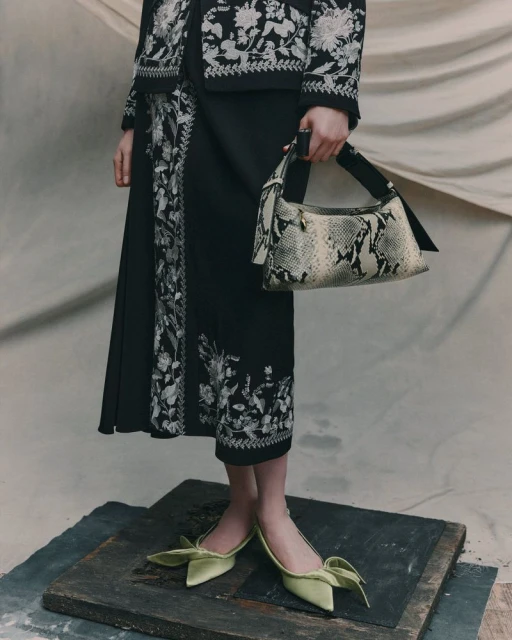Fashionable woman in black skirt and jacket with floral embroidery, holding a snakeskin handbag, and wearing green bow-tie heels.
