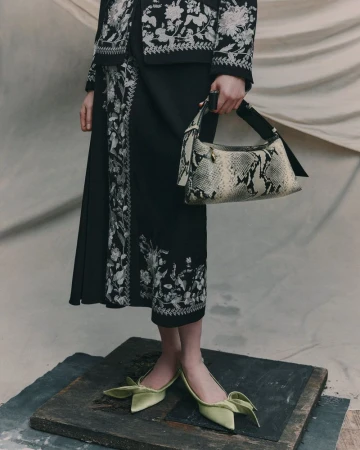 Fashionable woman in black skirt and jacket with floral embroidery, holding a snakeskin handbag, and wearing green bow-tie heels.