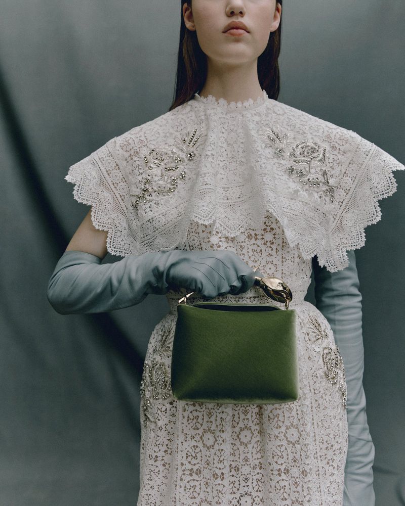 Young woman poses in a white lace dress and light blue gloves, holding a small green handbag against a muted gray background.