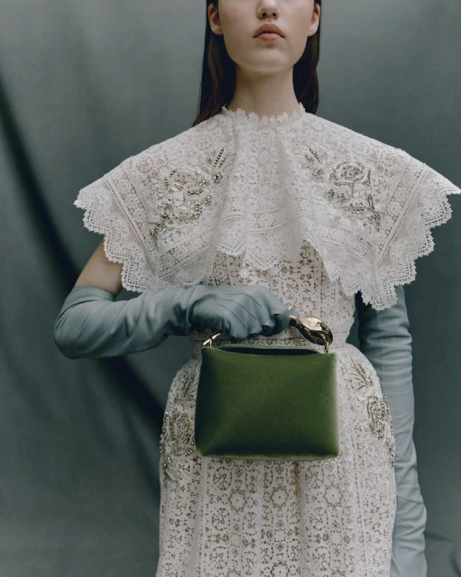 Young woman poses in a white lace dress and light blue gloves, holding a small green handbag against a muted gray background.
