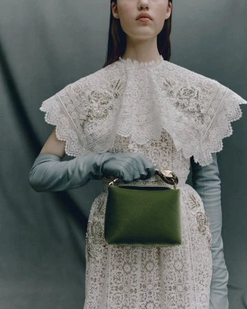 Young woman poses in a white lace dress and light blue gloves, holding a small green handbag against a muted gray background.