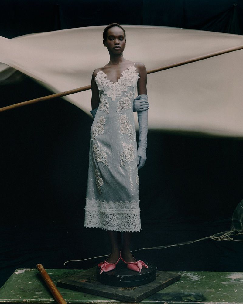 Elegant Black woman poses gracefully in a light blue dress with lace details, opera gloves, and pink shoes against a dark backdrop.