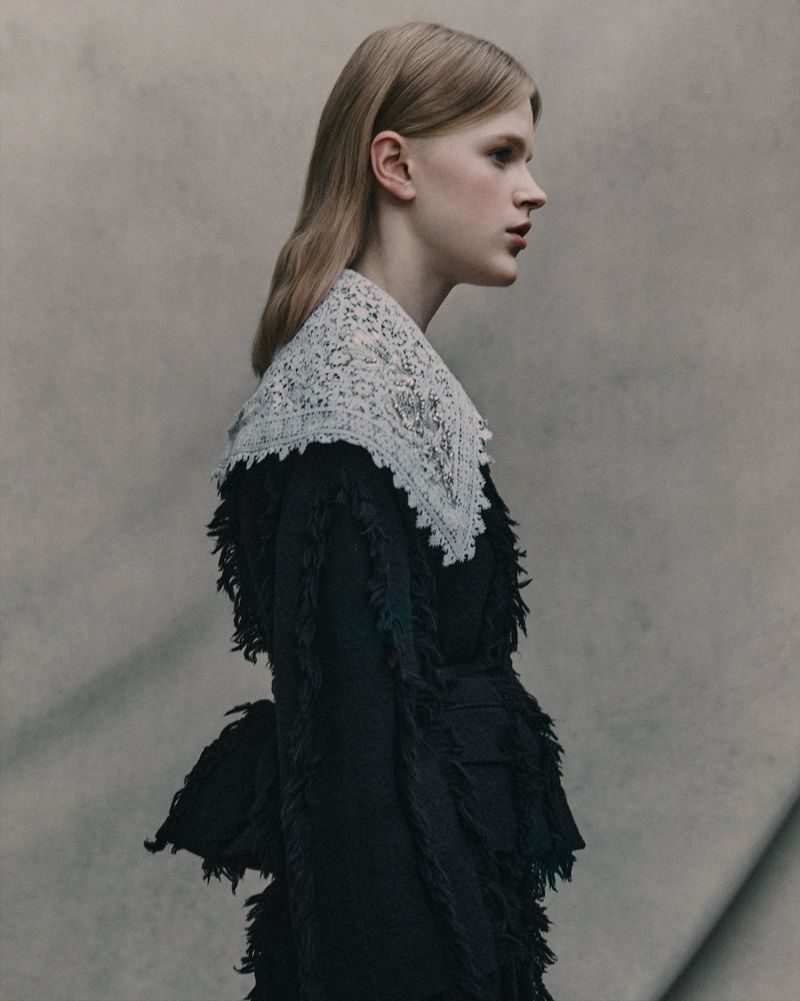 Profile shot of a blonde woman wearing a black textured jacket with a large white lace collar against a mottled gray backdrop.