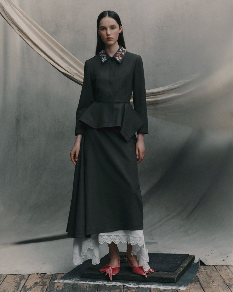 Elegant woman in a dark skirt and jacket with floral collar poses in front of a draped background, creating a studio fashion shot.