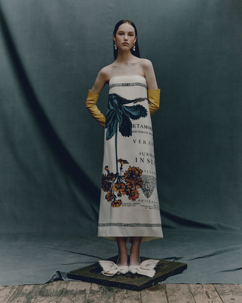 Elegant woman poses in a strapless dress with floral and text prints, complemented by long gloves, in a studio setting against a grey backdrop.