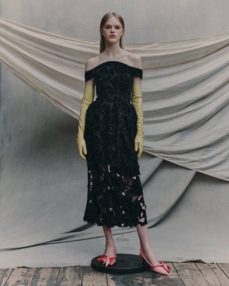 A woman models a black, off-the-shoulder dress with yellow gloves and pink shoes in a studio setting with draped fabric backdrop.