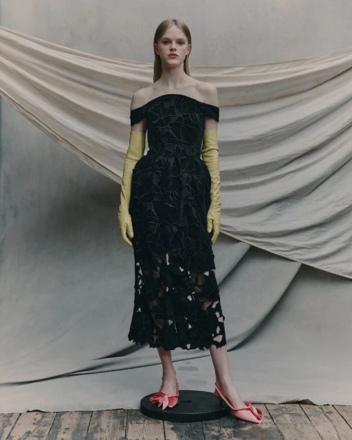 A woman models a black, off-the-shoulder dress with yellow gloves and pink shoes in a studio setting with draped fabric backdrop.