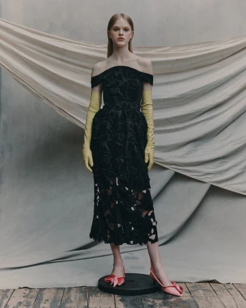 A woman models a black, off-the-shoulder dress with yellow gloves and pink shoes in a studio setting with draped fabric backdrop.