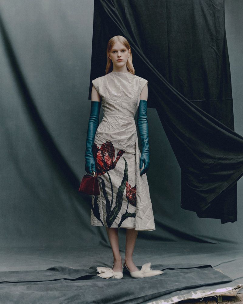 Full-length shot of an adult woman posing in a floral print dress, long gloves, and heels against a grey backdrop with a black drape.