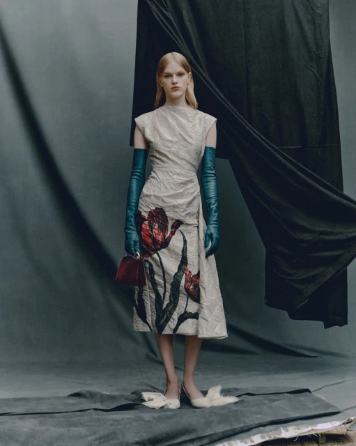 Full-length shot of an adult woman posing in a floral print dress, long gloves, and heels against a grey backdrop with a black drape.