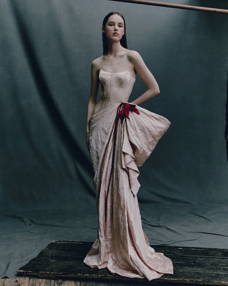 A woman poses in a light pink strapless gown with a large bow detail on a platform against a grey textured backdrop in a fashion shot.