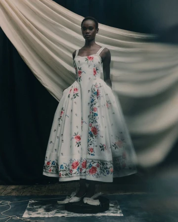 Adult Black woman posing in a full-skirted floral gown in a studio setting with a draped backdrop and worn flooring, fashion photography.