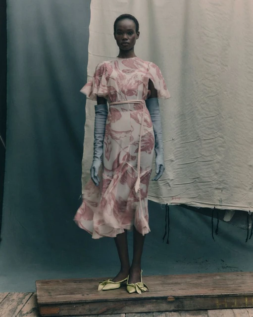 A full shot of an African American female model wearing a floral midi dress, light blue gloves, and yellow shoes on a wooden platform.