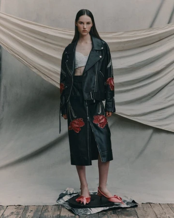 Full-length studio shot of a young adult woman in a black leather jacket, skirt, white top, and pink bow shoes against a beige backdrop.