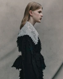 Model in profile wearing a black textured garment with a white lace collar against a grey studio background.